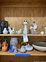 Shelf with white ceramic bird figurines, two Willow Tree figurines, small bowl, and assorted miniature vases and pitchers in pastel and neutral colors.