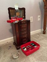 Photo of wooden jewelry box with mirror and multiple drawers, with red trays of costume jewelry including earrings and bracelet on floor beside it.