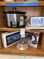 Cuisinart slow cooker on wooden shelf showing front controls and glass lid alongside boxed vaporizer, cordless drill, bird feeder, digital device, and recipe book visible.