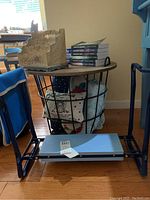 Gardening kneeling bench with blue metal frame and beige fabric tool pockets, mesh basket with blankets underneath, wooden organizer box on bench, sets of books on top and floor.