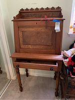 Front view of closed drop front desk showing decorative carved wood back rail and single drawer below.