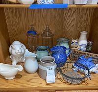 Overall view showing assorted sauce pitchers, decorative ceramic rabbit, turtle, glass jars, and small animal figurines arranged on wood shelf.