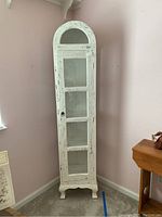 Front view of tall narrow shabby chic cabinet in distressed white finish with glass front door and shelves visible inside.