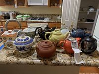 Photo of all nine teapots arranged on granite countertop showing variety of styles, colors, and shapes