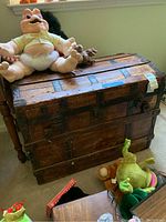 Front and top view of vintage wooden chest with toys placed on top and around it, showing dark metal reinforcements and weathered wood surface.