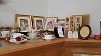 Photo showing a variety of vintage cups, saucers, decorative plates, bowls, glassware, and framed art pieces on a wooden surface.