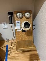 Full front view of the oak wall-mounted vintage telephone showing the wood body, metal bells, handset, and speaker area.