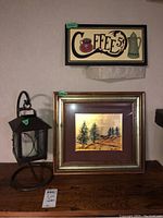 Photo showing the coffee-themed sign wall art above the framed pine tree artwork and metal lantern on a wooden surface.
