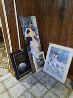 Four framed and mounted prints leaning against wood wall and door, includes floral prints, children with geese print, and the Firefighter's Prayer print on board.