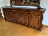 Image showing the front and side view of the long wooden buffet cabinet with ornate carved doors and drawers, wood grain top, standing on wood floor, with visible wear and some damage on top rear edge