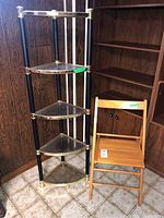 Full view of corner cabinet with four glass shelves and wood folding chair in front, side angle