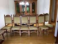 Six carved wooden dining chairs with upholstered seats and backs, arranged in a row in front of a dining cabinet.