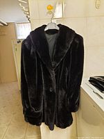 Photo showing front view of black faux fur coat hanging on a hanger, highlighting the coat’s texture and button closure.
