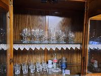 Two shelves inside a wooden glass cabinet featuring an assortment of glassware including sherry glasses, short shot glasses, a Coca-Cola bottle, and other small glass containers.