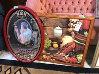 Photo showing both the Coca-Cola oval mirror with a red frame and the framed kitchen-themed print side by side on the floor with furniture in the background.
