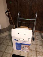 View of walker, three canes, and boxed raised toilet seat against wall and floor.