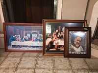 Three framed religious wall arts on tile floor: large Last Supper print, medium framed biblical scene, smaller framed portrait of Padre Pio.