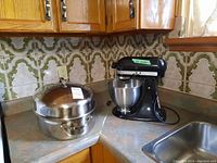 Stainless steel roaster pot with domed lid and KitchenAid black stand mixer with bowl on kitchen counter near sink.