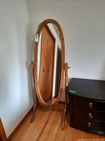 View of full standing oval mirror on wooden swivel base, next to dark wood dresser against a white wall and wooden door