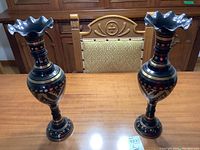 Two matching handcrafted metal vases approximately 18 inches tall placed on a wooden table in front of a carved wooden chair.