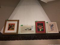 Four framed cross stitch wall art pieces arranged side by side showing variety of subjects: bird, rose with text, two floral designs.