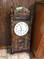 Full view of the carved wooden Tulxis wall clock showing the clock face, carved top, side columns and pendulum window.