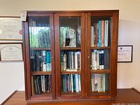 Front view showing wooden bookcase with three glass doors containing books, brass knobs on doors.