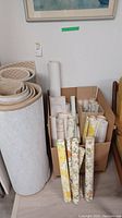 Photo showing multiple wallpaper rolls standing upright with floral and plain patterns, two large rolled carpets beside the wallpaper.