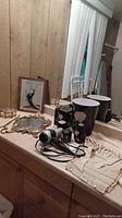 Overview of vintage bathroom vanity items including ceramic containers, mirrors, framed figurine artwork, and vintage corset on countertop.