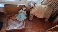 Photo showing porcelain doll in blue dress inside an open cardboard box, plastic bubble wrap present in box, standing next to large woolly plush sheep dog on wooden floor.
