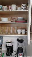 Cabinet shelf with mugs on top, Corelli dishes in middle, clear glasses and additional mugs below; black coffee maker and Hamilton Beach blender on counter below the cabinet.