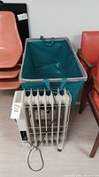 Rolling laundry cart with green fabric basket and Delonghi oil heater in front of chairs.