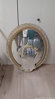 Large oval gilded mirror leaning against a wall on floor, showing its full frame and reflective surface.