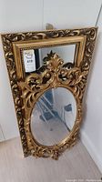 View of both gilded mirrors leaning against a wall, showing ornate frames and inset oval mirror on the larger piece.