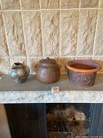 Three pottery vessels aligned on a stone hearth against a tiled wall: the smoky signed vase on the left, the lidded stamped pot in the center, and the open clay vessel on the right.