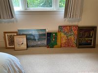 Overview of six framed art prints and one unframed art piece arranged on floor against wall near window