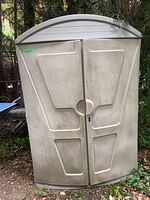 Front closed view of beige/grey outdoor plastic storage unit with a curved top and double doors showing some dirt and wear.