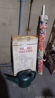 Boxed West Bend Redwood outdoor grill table alongside rolled patio umbrella and green watering can.