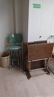 Vintage green vinyl folding stool with chrome metal legs next to beige ceramic crock and metal TV trays and folding wooden table.