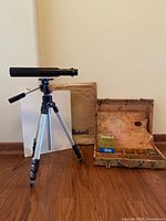 Spotting scope on full tripod, painters box, bag and blank canvases on wood floor.