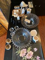 Overview of table with two large glass serving bowls, three small clear glass bowls, floral ceramic trays, dinner place cards, coasters, and other ceramic items including the matchbox holder.