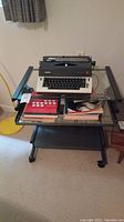 Olympia electric typewriter placed on rolling glass desk with additional items on lower shelf.