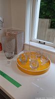 6 clear crystal snifters displayed on a bright yellow round serving tray next to Nachtmann vase with giraffe etching.