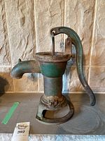 Full view of the vintage hand pump showing its cast iron body, lever handle and outlet spout with paint chipped and rusted areas.