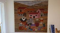 Full view of the tapestry hung on a wall, showing village houses and people in traditional dress with hats and colorful baskets.