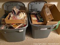 Two large plastic storage bins filled with various candles and other items, showing an assortment of taper candles mostly wrapped in plastic in one bin and boxes in the other.