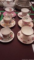 View of six assorted vintage china cups and saucers from six different prestigious brands arranged on a table, showing floral and gold trim patterns.