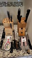 Two wooden knife blocks on granite countertop with multiple knives inserted and laid out in front, along with a decorative spoon rest and kitchen sponge