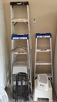 Photo showing full height 6 ft aluminum ladder, two 2 ft aluminum step ladders, white plastic step stool and black metal folding dolly.