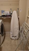 Photo showing an ironing board with a white cover standing upright, some metal drying racks leaning against wall, and part of a washer or dryer.
